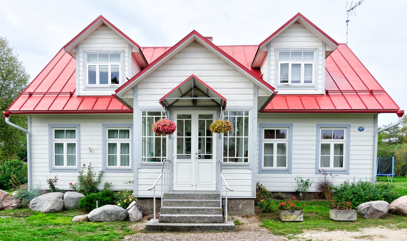 White clapboard cottage with a bright red metal roof, dormer windows, and hanging flower baskets by the front door